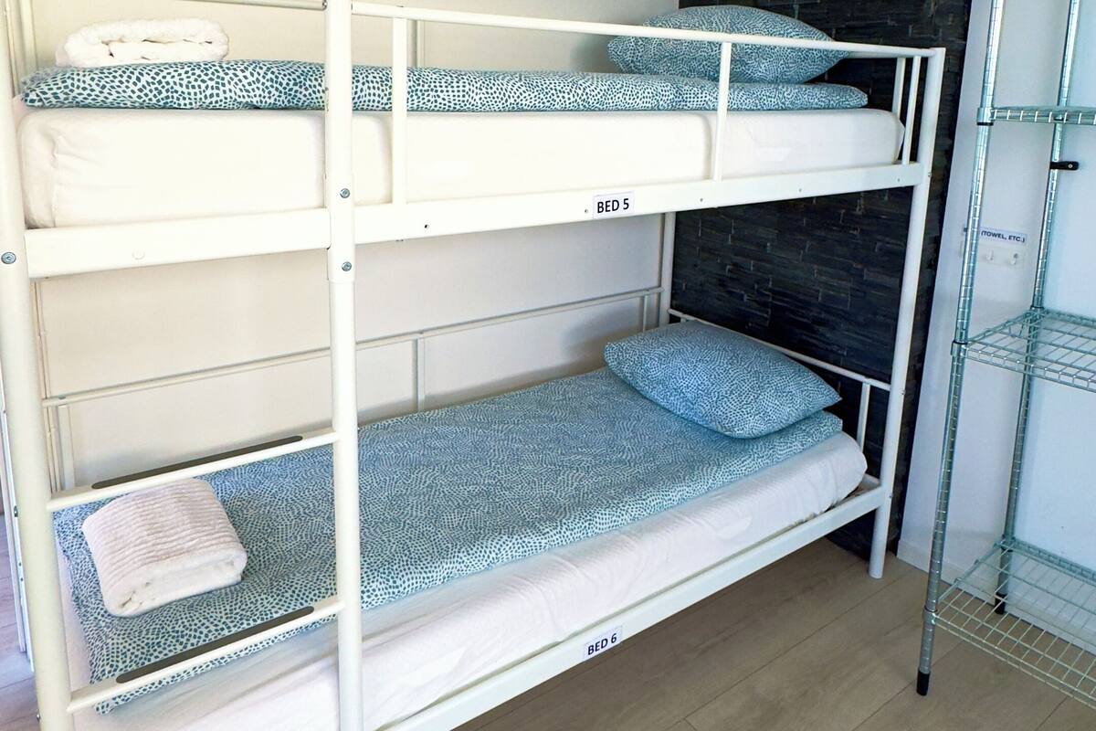 A bunk bed with three levels, labeled BED 5 and BED 6, in a room with wooden flooring and a dark brick wall.