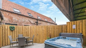 Un patio trasero con jacuzzi, sillas de mimbre, cerca de madera y un edificio de ladrillo con techo de tejas.