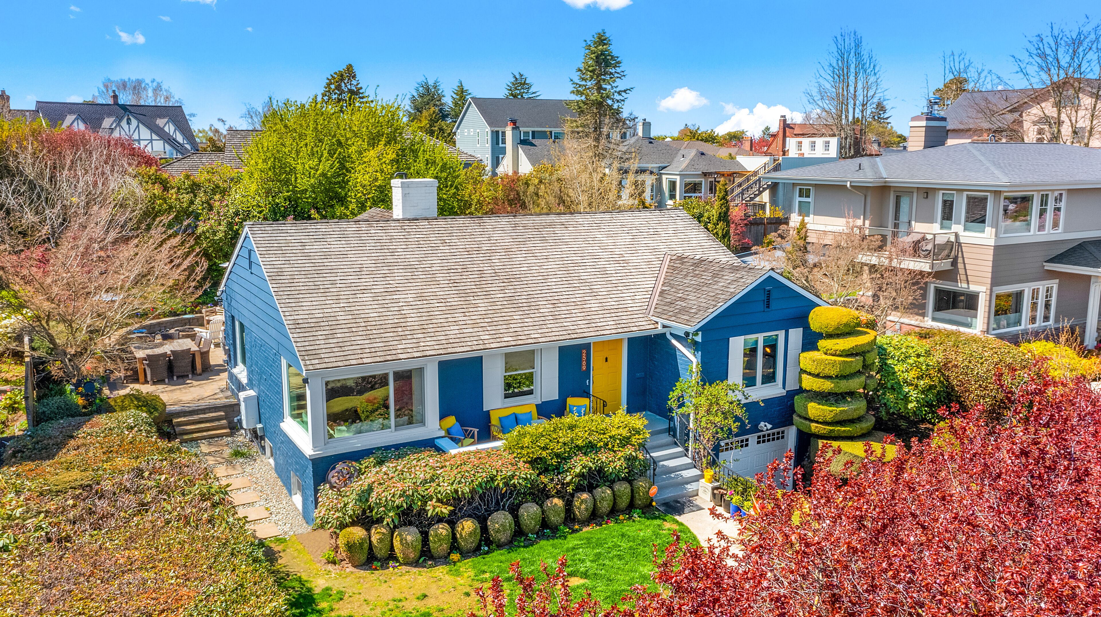 A blue house with a grey roof, surrounded by lush greenery and other houses.