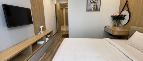 A modern hotel room with a flat-screen TV, a bedside table with a lamp, a wooden nightstand, and a bed with white bedding.