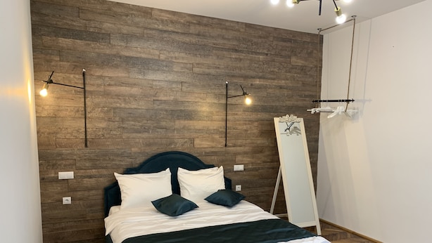 A bedroom with a wooden wall, a bed with white and dark bedding, and a standing mirror. - 1793 Margarethengasse Guesthouse (Sibiu)