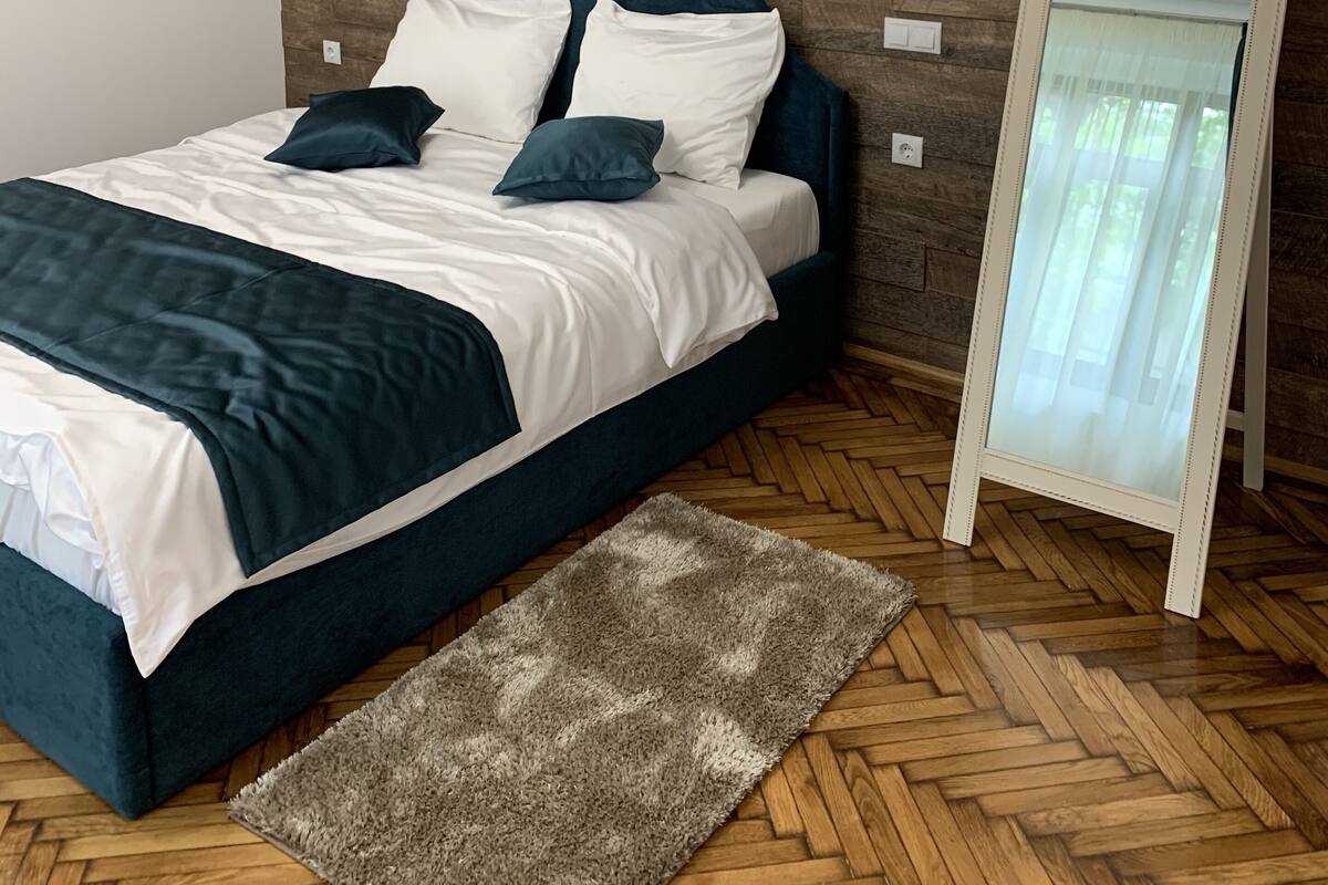 A modern bedroom with a wooden wall, a bed with white and dark blue bedding, a mirror, and a herringbone-patterned floor.