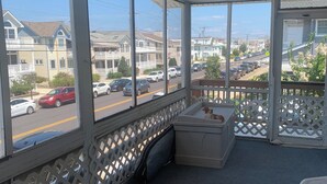 Un espace extérieur abrité, donnant sur une rue résidentielle, avec un banc blanc et une petite table agrémentée d’un coussin.