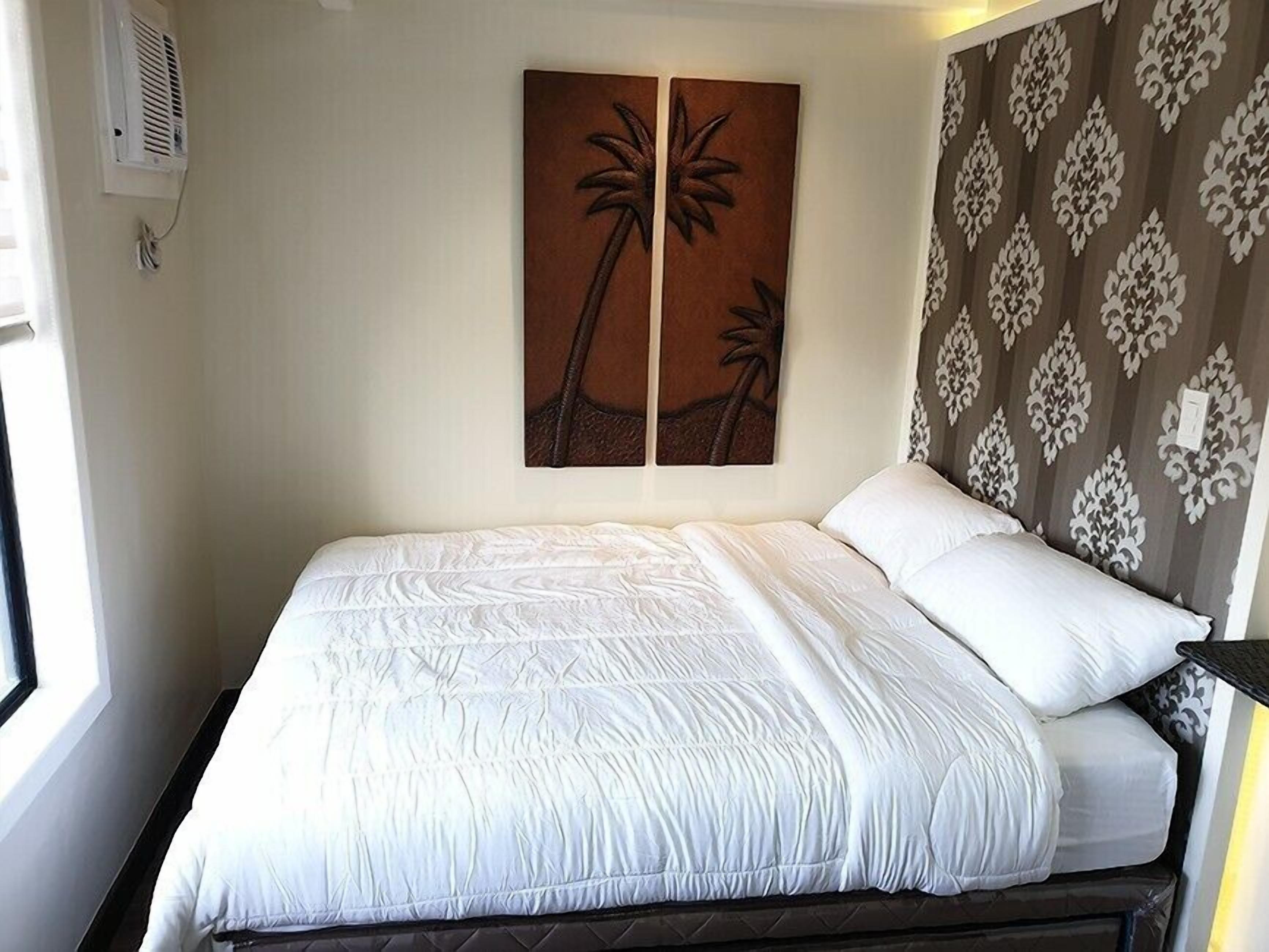 A bed with white bedding, a wall-mounted air vent, and a decorative wall panel with palm tree designs.