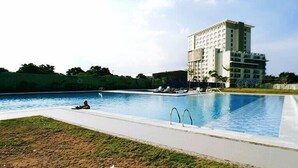 A swimming pool with lounge chairs and a person relaxing by the pool. A large building with multiple floors is visible in the background. - Soltana Nature Residence (Lapu-Lapu)
