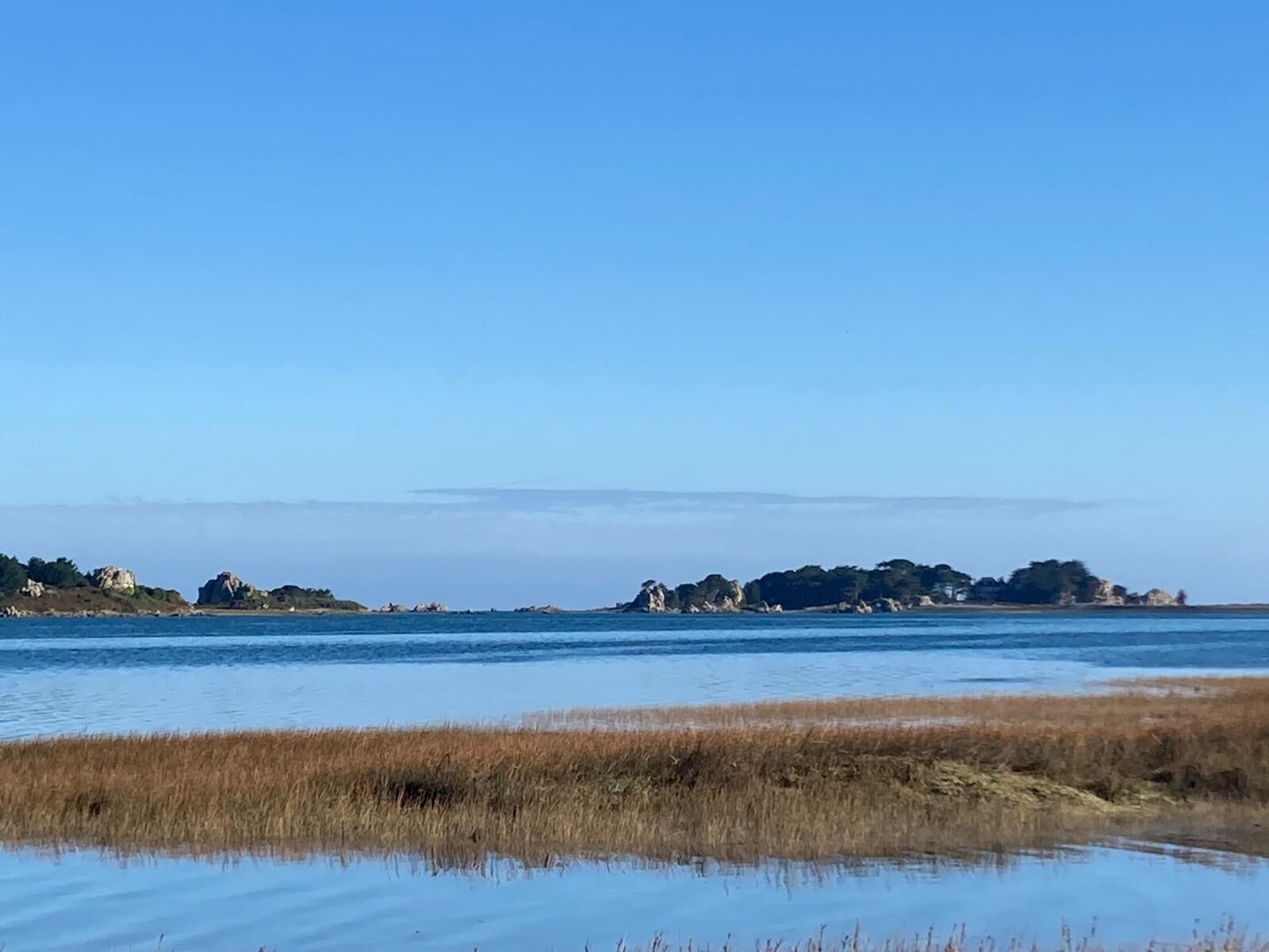 Un paysage côtier caractérisé par un ciel dégagé, une eau calme et des formations rocheuses lointaines.
