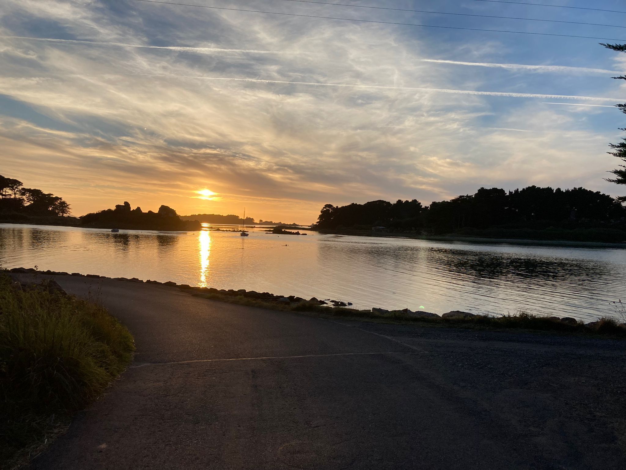 Ein Sonnenuntergang über einem ruhigen Fluss mit Booten, eine Straße im Vordergrund und Bäume im Hintergrund.