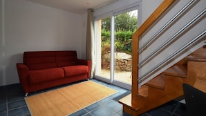 A room with a red sofa, a wooden staircase, and a view of a garden through a glass door. - Les Palmiers D'armor (Penvénan)