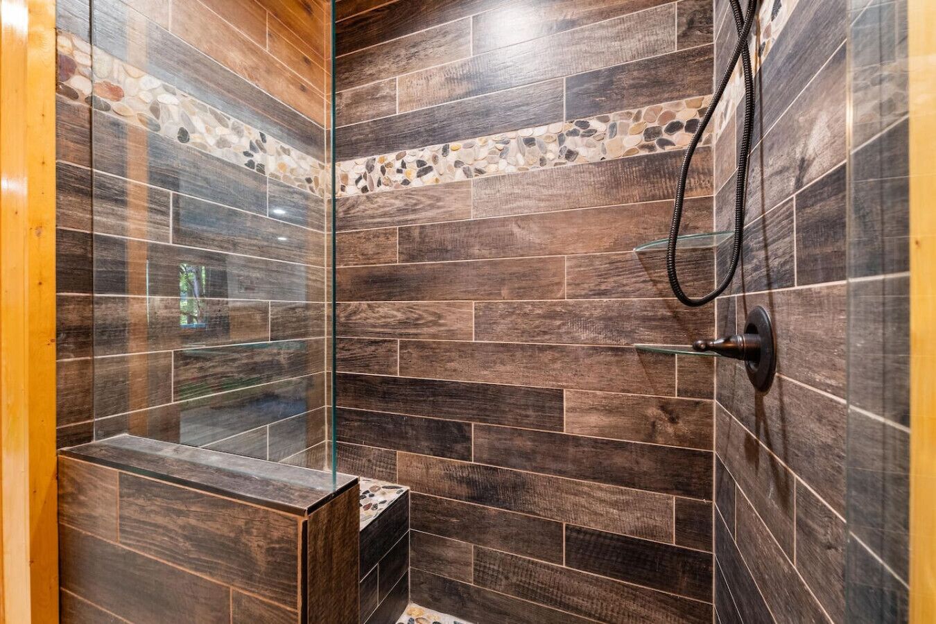 Ducha con azulejos oscuros y puerta de vidrio, cabezal de ducha visible y un estante pequeño.