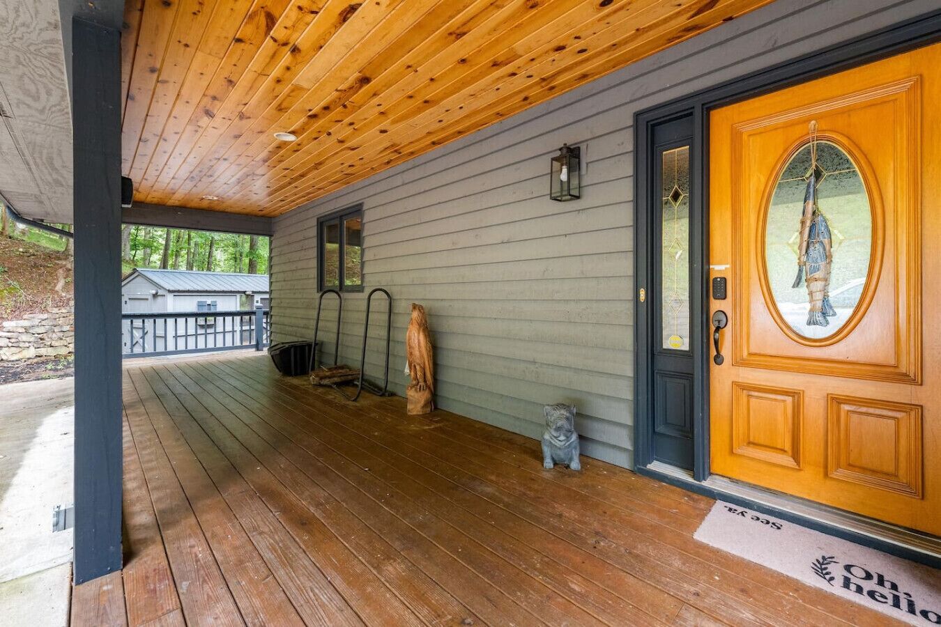 Un portico in legno con una casa rivestita di grigio, una porta in legno con un pannello di vetro decorativo e una piccola statuetta di un gatto.
