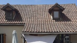 Una casa con techo de tejas, un ventanal pequeño y un toldo blanco triangular sobre una mesa y sillas.