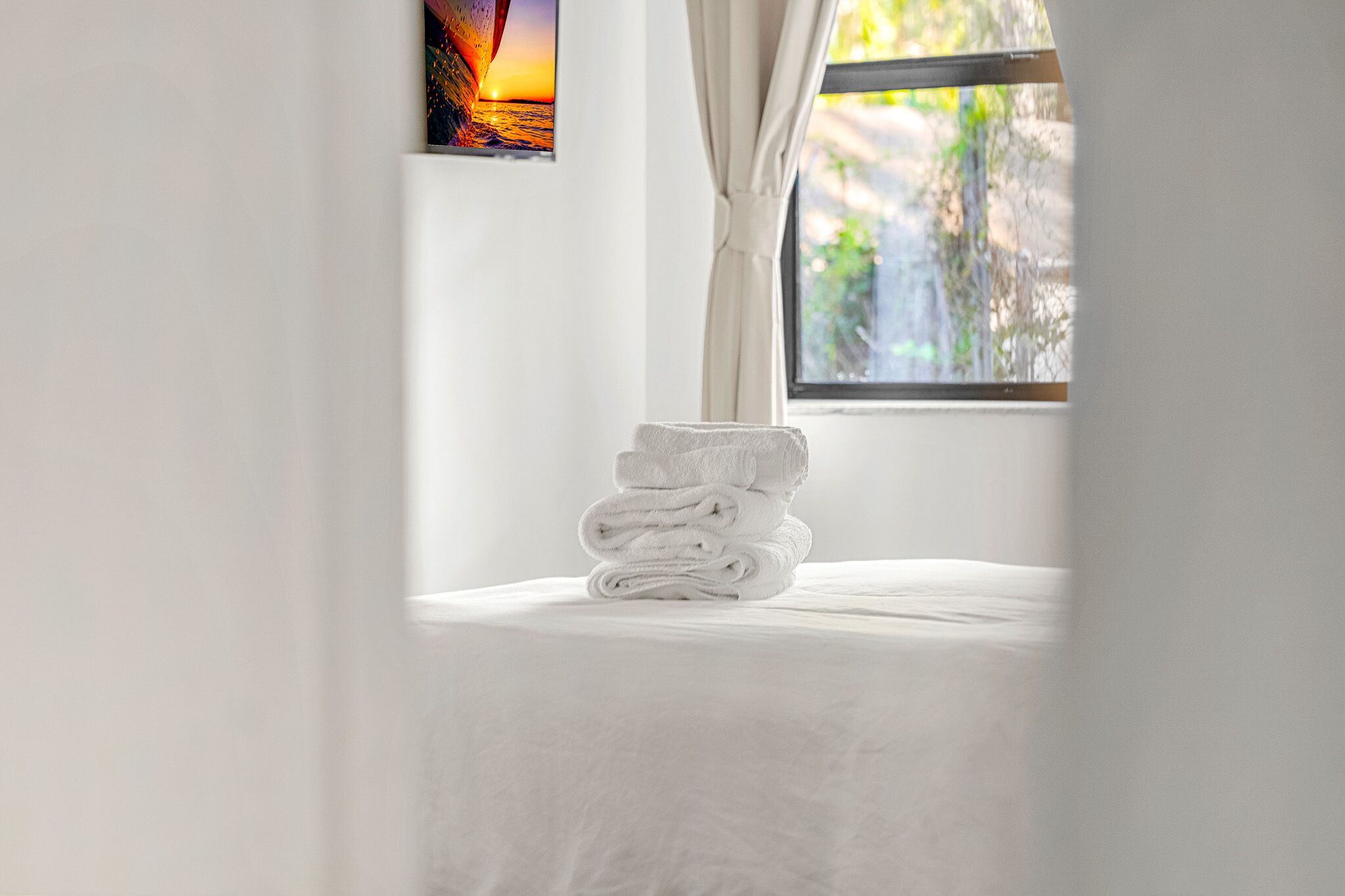 Una cama bien hecha con una pila de toallas blancas, una ventana con vistas a la vegetación y un cuadro en la pared.