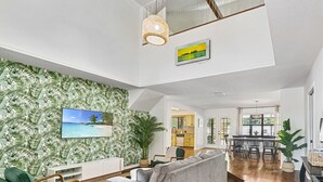 Ein geräumiges Wohnzimmer mit einem grauen Sofa, einem grünen Sessel und einem Holztisch. An den Wänden befinden sich ein großes Wandbild mit einer tropischen Szene und ein gerahmtes Bild.