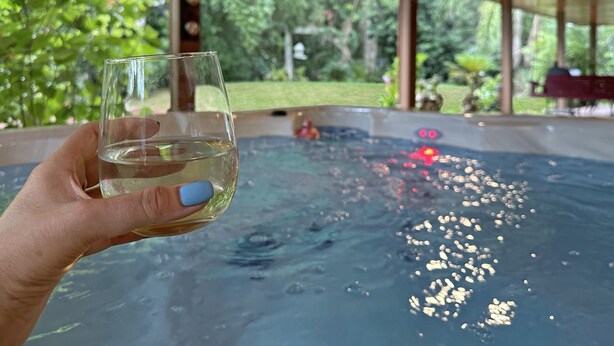 Eine Hand hält ein Glas Wasser in der Nähe eines Whirlpools mit Blick auf einen Garten.