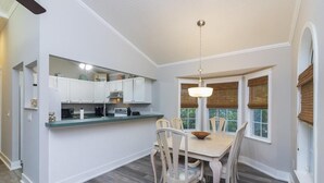 A dining area with a wooden table and chairs, a kitchen with white cabinets, and a window with a view of greenery. - Spacious Beach Home - 2 En-suites - Elevator (Saint Augustine)