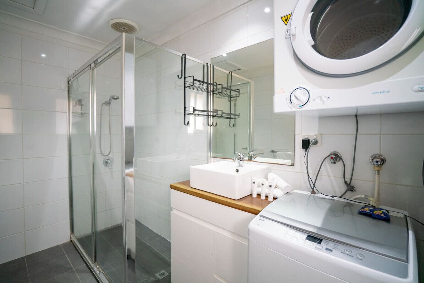 A modern bathroom with a glass shower enclosure, a white sink with a wooden countertop, and a built-in washing machine.