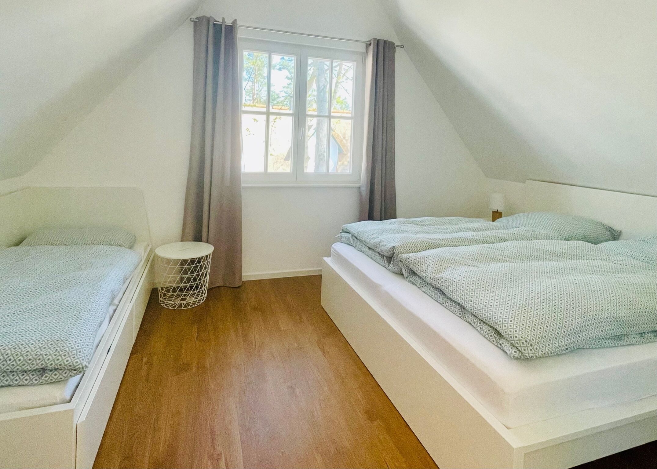 A bedroom with two beds, a window with curtains, a small round table, and a wooden floor.