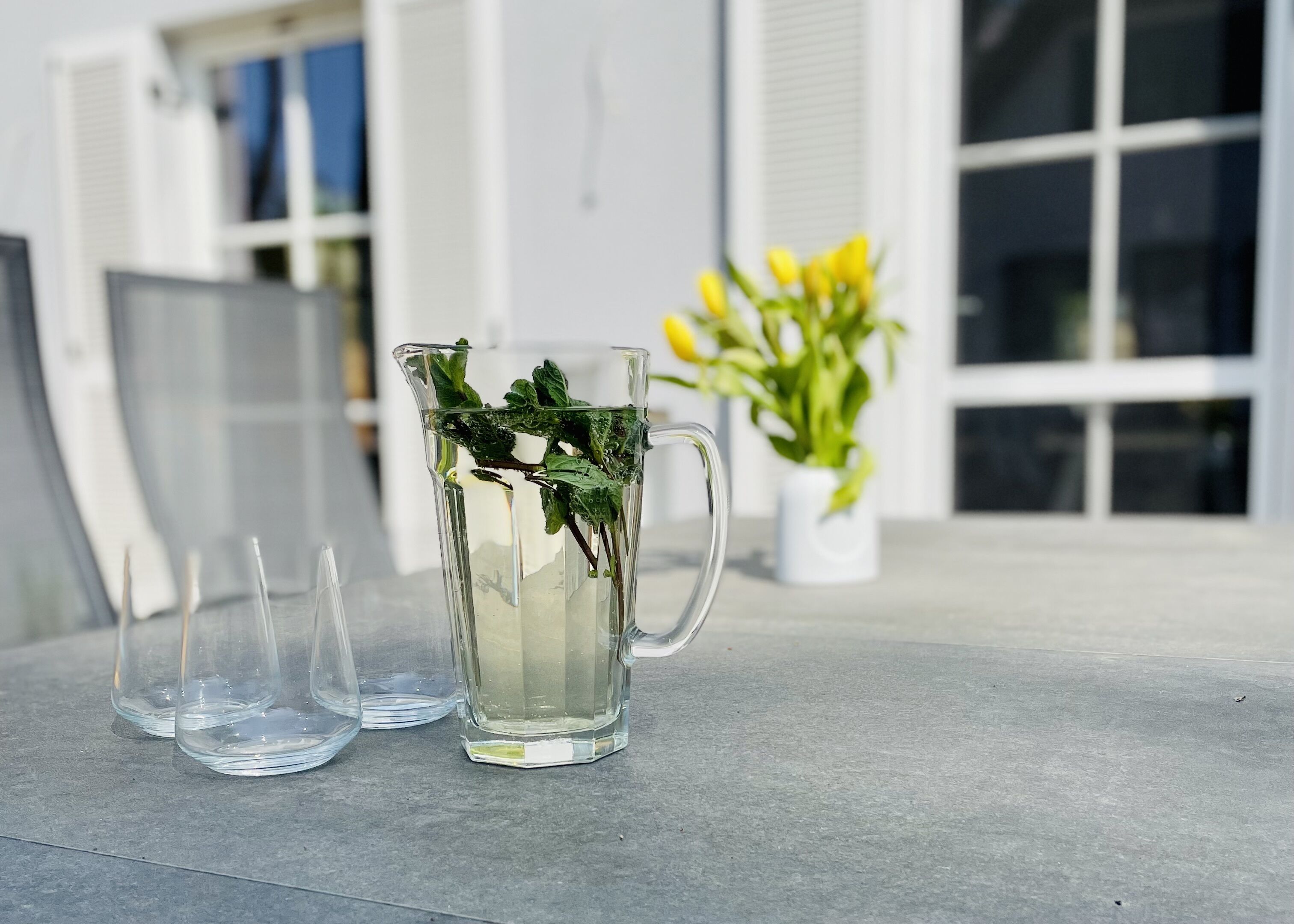 A clear glass pitcher with mint leaves on a table, accompanied by two empty glasses, in front of a building with a window and a potted plant with yellow flowers.
