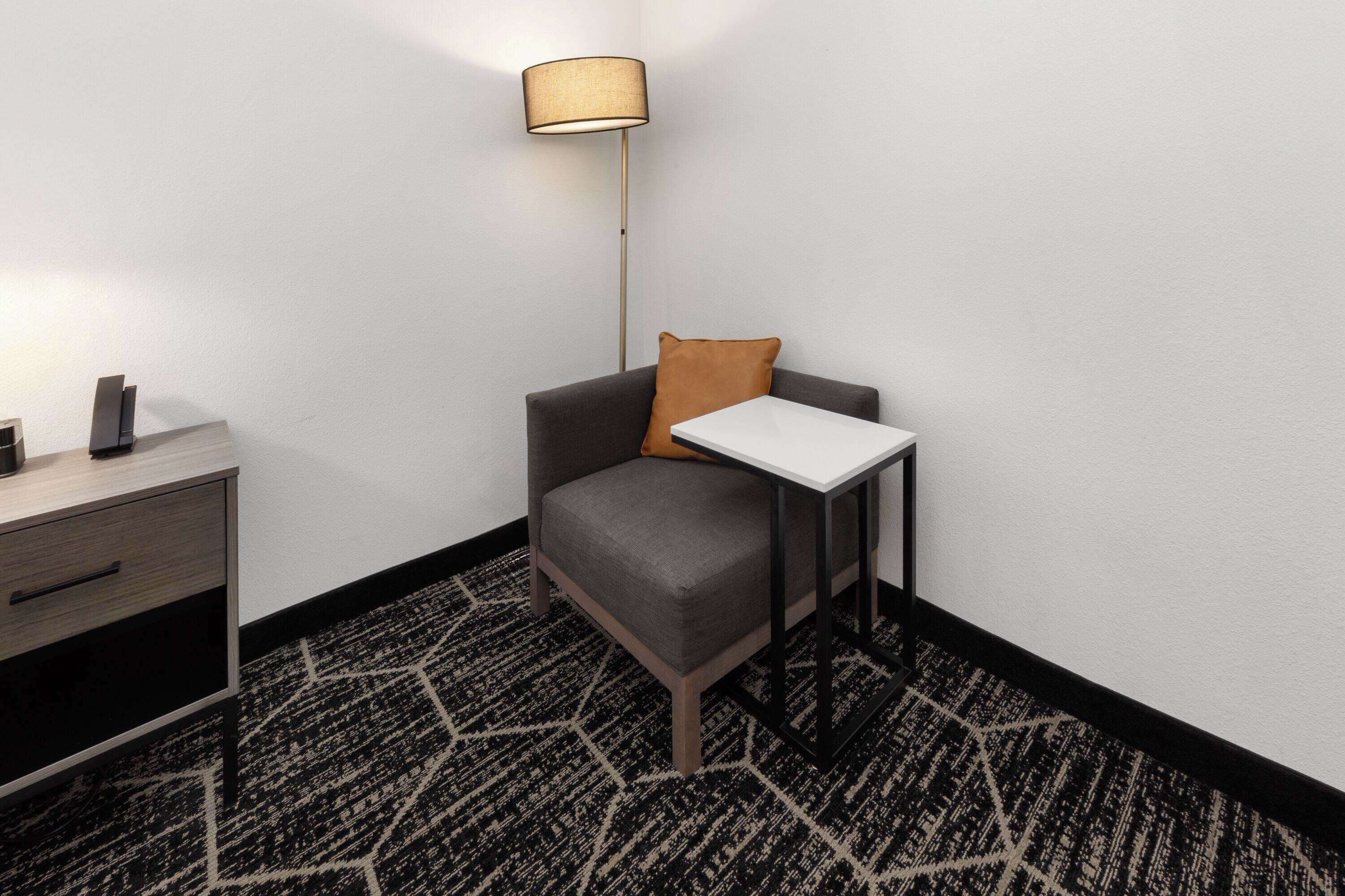 Una habitación de esquina con un sillón gris, una mesita blanca pequeña, una lámpara y una alfombra estampada.