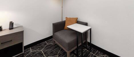 Una habitación de esquina con un sillón gris, una mesita blanca pequeña, una lámpara y una alfombra estampada.
