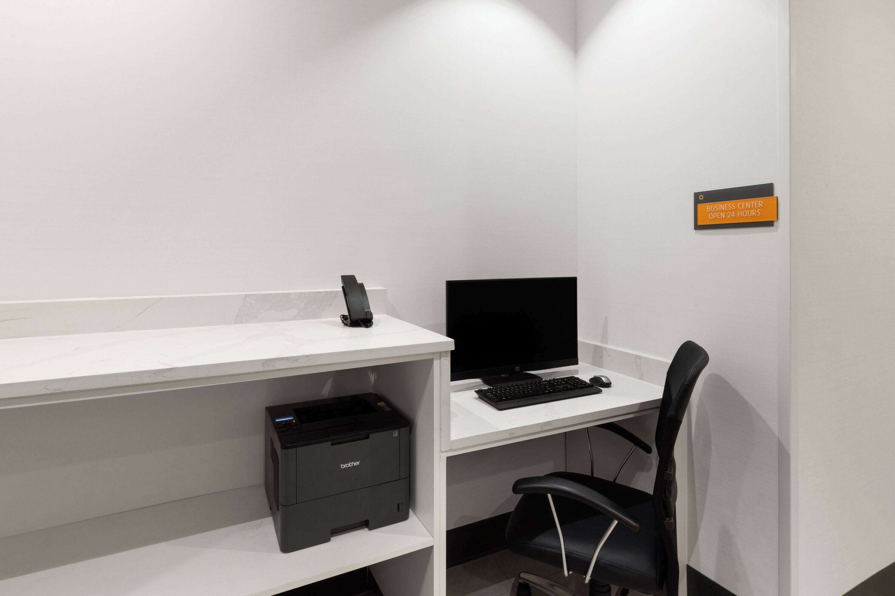 A clean, modern office space with a white desk, black office chair, and a printer. A sign indicates the business center is open 24 hours.