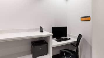 A clean, modern office space with a white desk, black office chair, and a printer. A sign indicates the business center is open 24 hours.