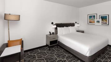 Quarto de hotel com cama, mesa de cabeceira, luminária, poltrona e pequena mesa.
