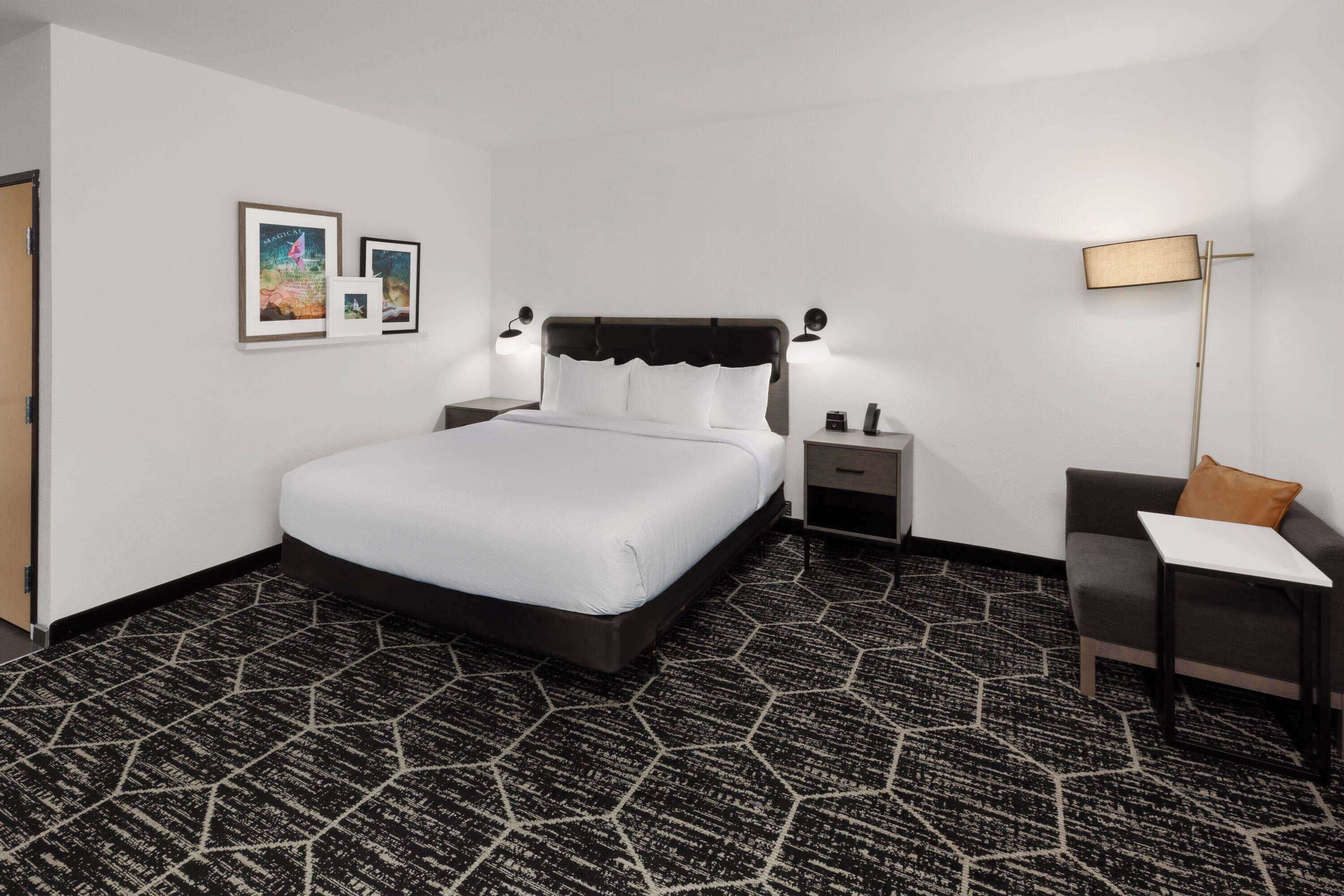 Une chambre d’hôtel avec un grand lit, une table de chevet, une commode, un bureau, une chaise et une lampe sur pied.