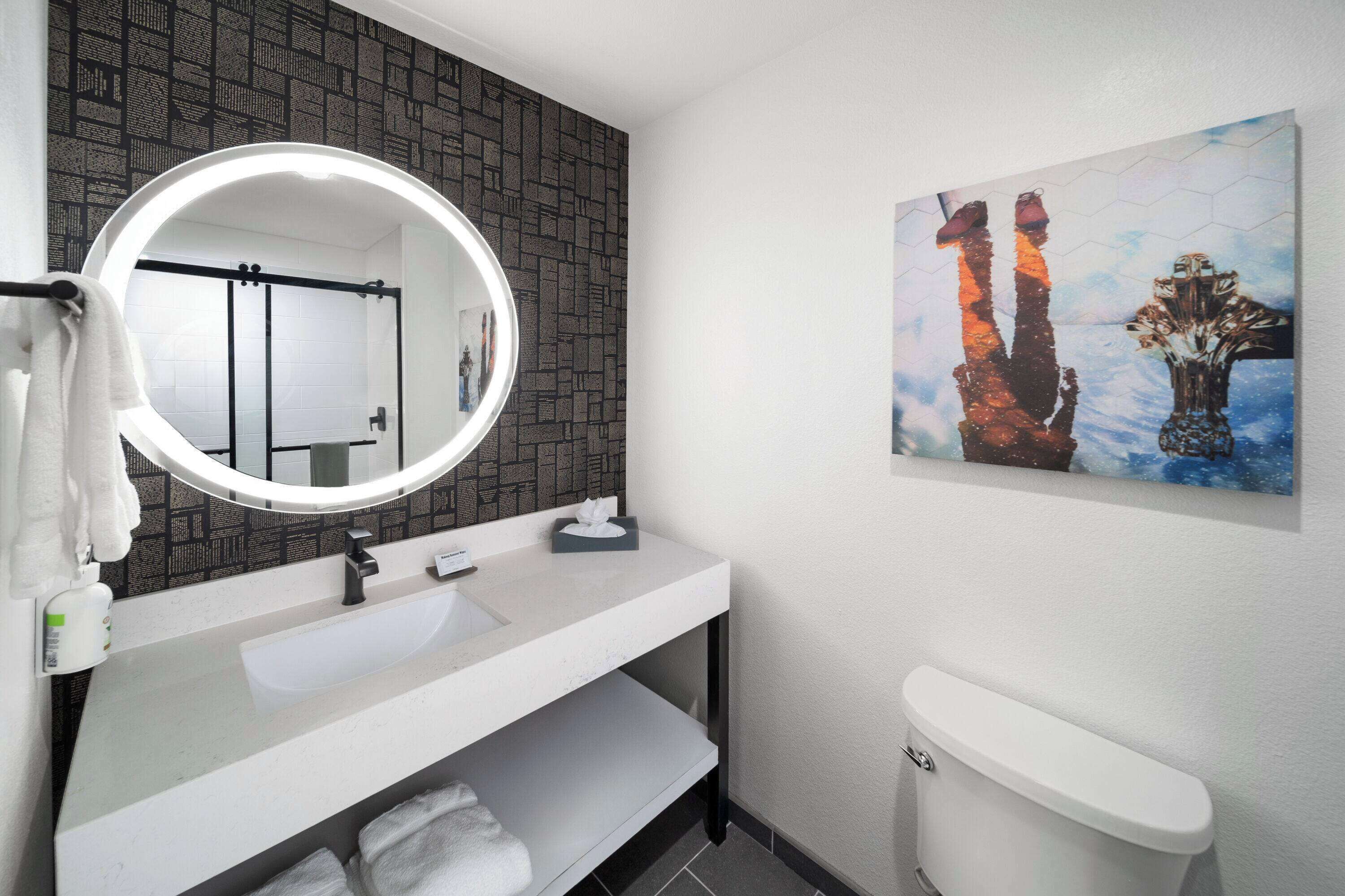 Un baño con un lavamanos blanco, un espejo redondo, un inodoro y una obra de arte en la pared.