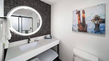 Un baño con un lavamanos blanco, un espejo redondo, un inodoro y una obra de arte en la pared.