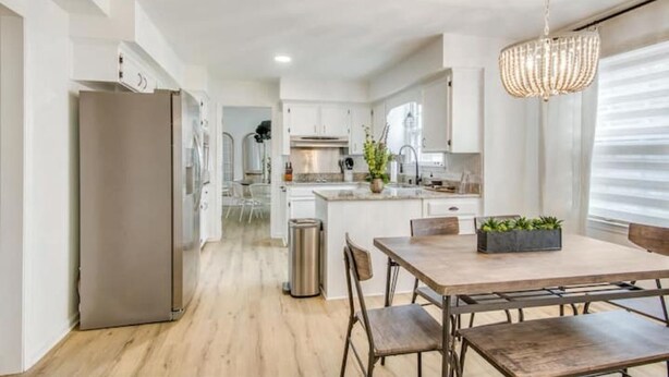 Una cocina moderna con armarios blancos, un refrigerador de acero inoxidable y una zona de comedor con una mesa y sillas de madera.