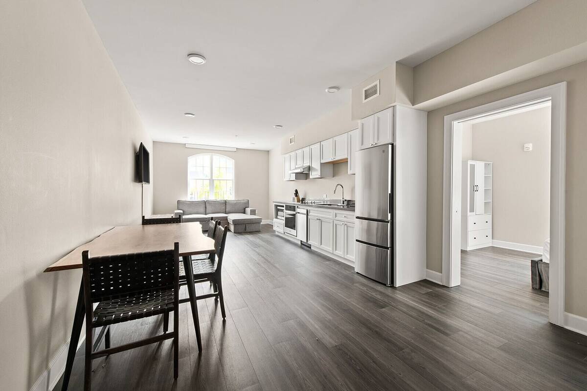 A modern kitchen with white cabinets, a stainless steel refrigerator, and a dining area with a wooden table and chairs.