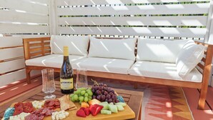 Una mesa de madera con una tabla de embutidos, una botella de vino y vasos en un patio con un banco acolchado blanco y una alfombra estampada.