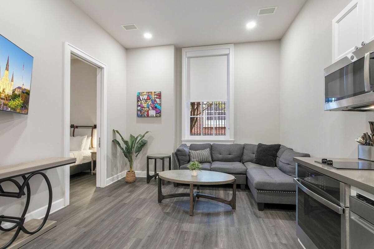 A modern living room with a gray sectional sofa, a round coffee table, a dining table with chairs, a large window with a view, and built-in appliances.