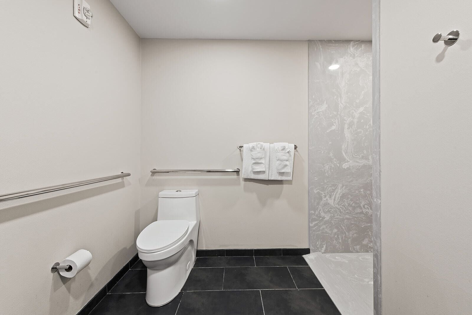A small, modern bathroom with a toilet, a handrail, a towel rack, and a roll of toilet paper.