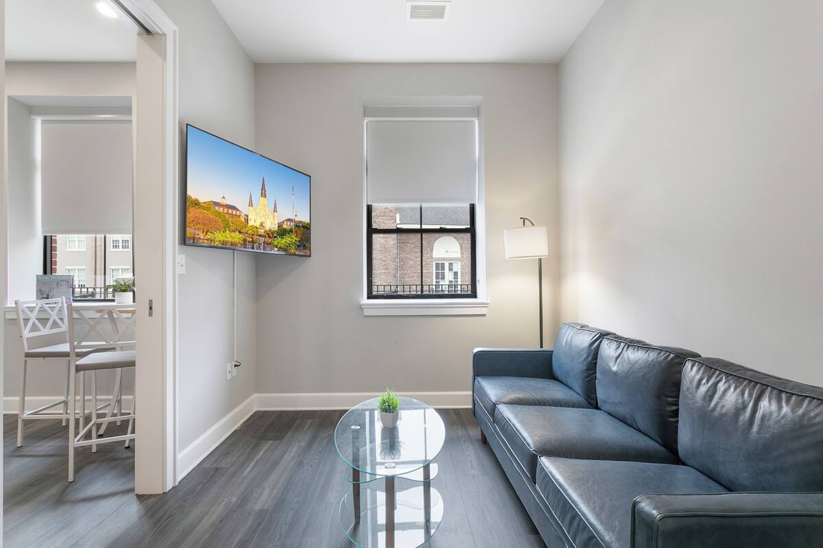 A modern living room with a glass coffee table, a black leather sofa, a flat-screen TV mounted on the wall, and a window with a view of a building.