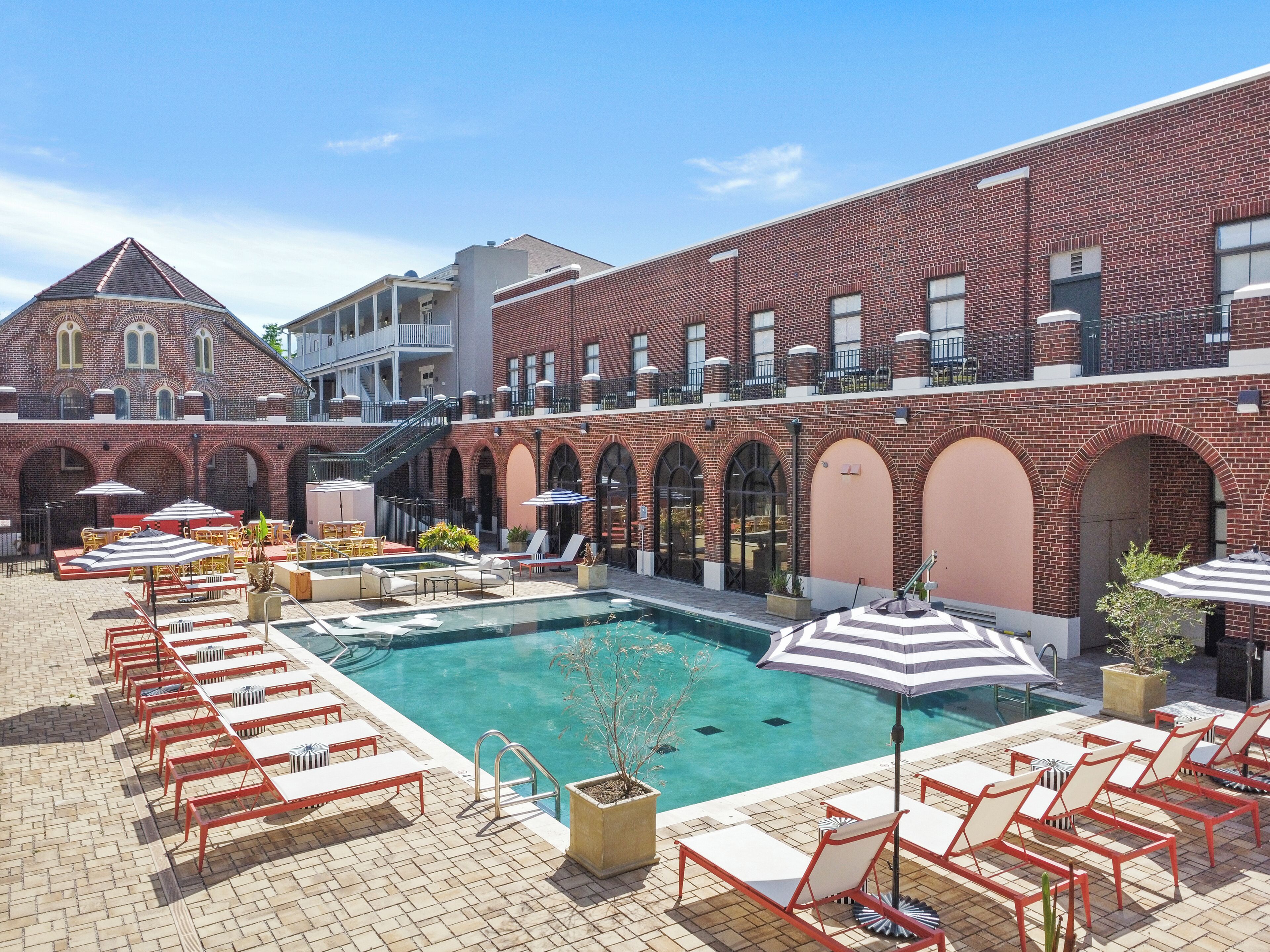 A rooftop pool area with lounge chairs and umbrellas, surrounded by brick buildings with arched windows and a staircase.