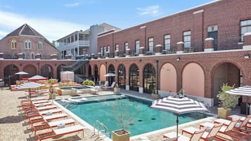 A rooftop pool area with lounge chairs and umbrellas, surrounded by brick buildings with arched windows and a staircase.