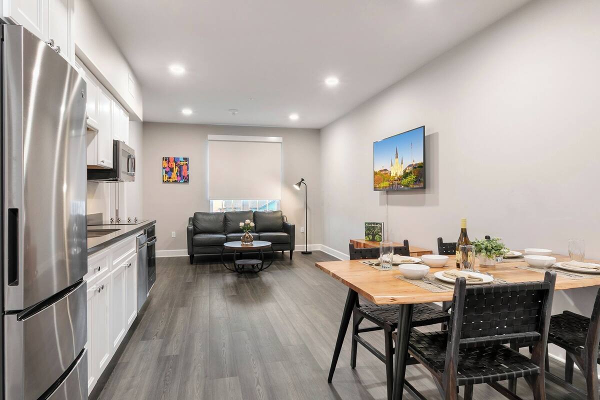 A modern kitchen with stainless steel appliances, a dining area with a wooden table and black chairs, and a black leather sofa in the living area.