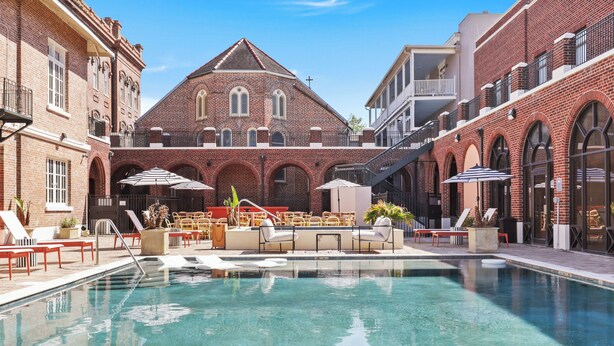 Zona de piscina con sillones de playa y sombrillas, rodeada de edificios de ladrillo con ventanas arqueadas y una imponente construcción que recuerda a una iglesia al fondo.