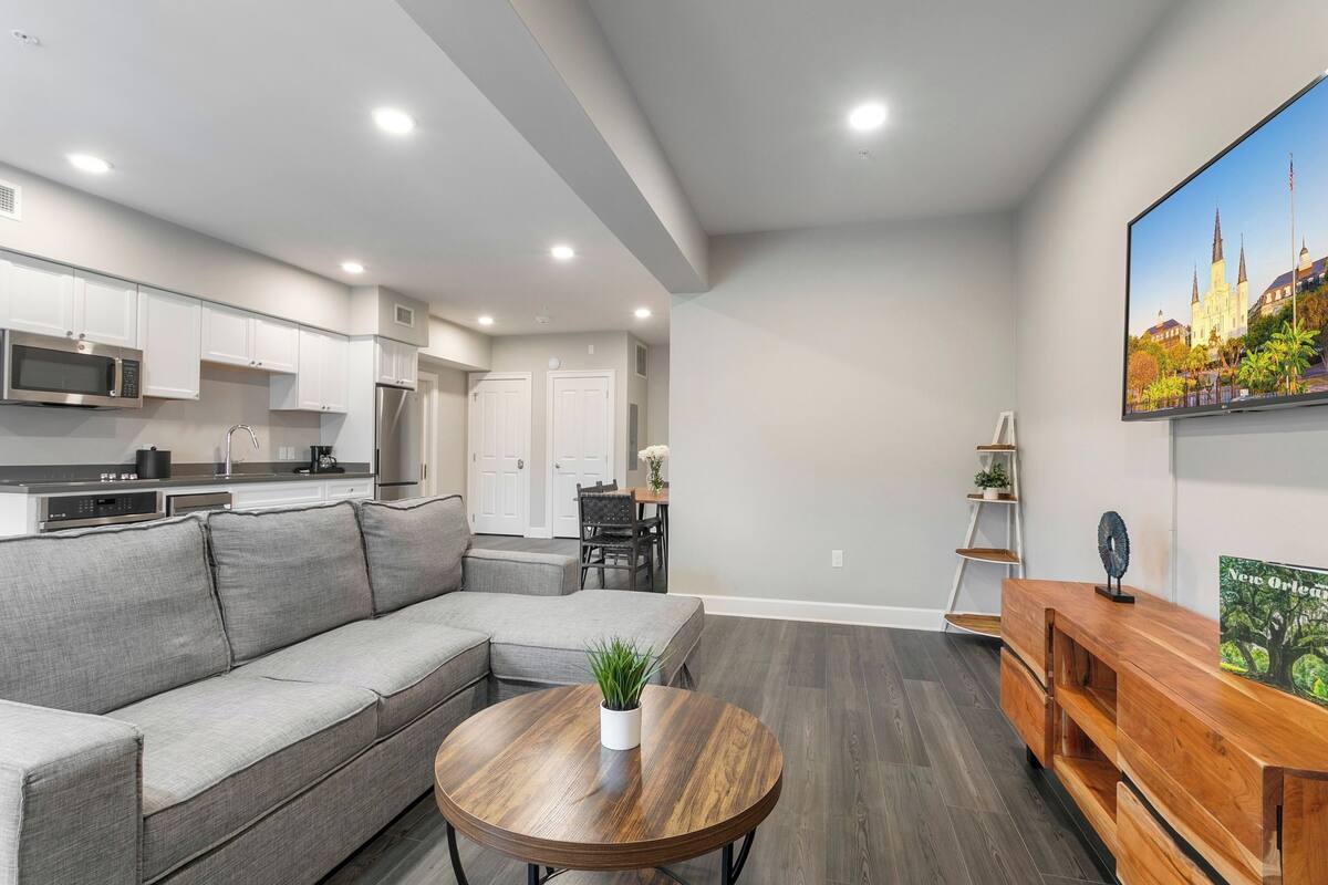 A modern kitchen with white cabinets, a gray sectional sofa, a round wooden coffee table with a plant, and a dining area with a white table and chairs.