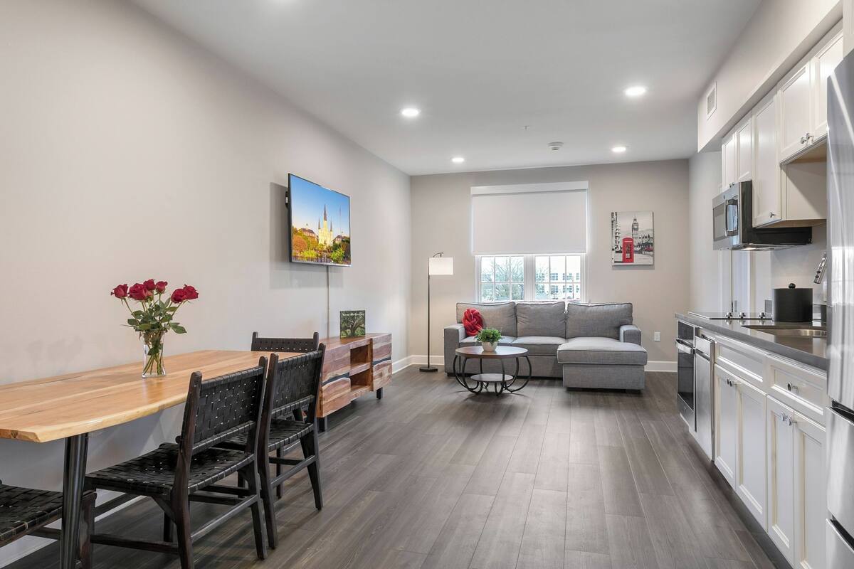 A modern kitchen with white cabinets and stainless steel appliances, a dining area with a wooden table and chairs, and a living area with a grey sofa and a small round table.