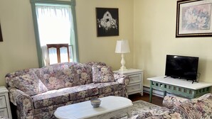 Una sala de estar con sofás de estampado floral, una mesa de centro blanca, un televisor sobre un mueble y obras de arte enmarcadas en las paredes.