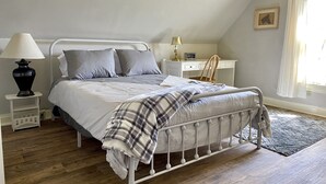 Un dormitorio con estructura de cama metálica, mesita de noche con lámpara, escritorio con silla y ventana con cortinas.