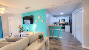 A modern living room with a teal accent wall, a flat-screen TV, and a kitchen area with a microwave and stove. - Refreshed 3BR condo- pool/patio/tennis - 1st Floor (Four Corners)