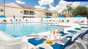 A pool area with lounge chairs and umbrellas, a pool with a clear blue water, and a building with a light-colored facade in the background. - Refreshed 3BR condo- pool/patio/tennis - 1st Floor (Four Corners)