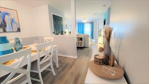 A modern dining area with white furniture, a wooden table with a decorative item, and a view into a living room with a blue couch and curtains. - Refreshed 3BR condo- pool/patio/tennis - 1st Floor (Four Corners)