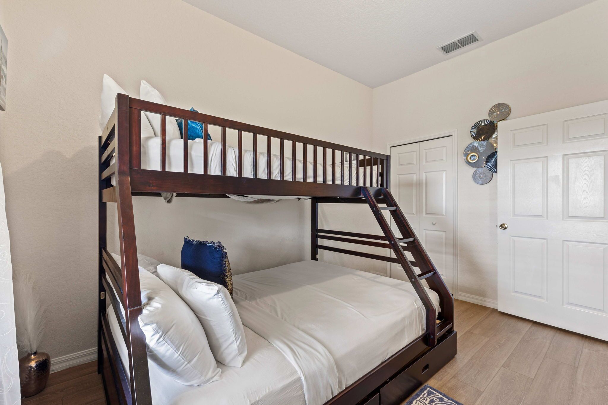 Een stapelbed met houten frame, wit beddengoed en een blauw kussen. Er is een witte deur met decoratieve platen aan de muur.
