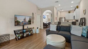 Ein Wohnzimmer mit einer Couch, Hocker, Fernseher und einem Essbereich mit Stühlen und einem Tisch.
