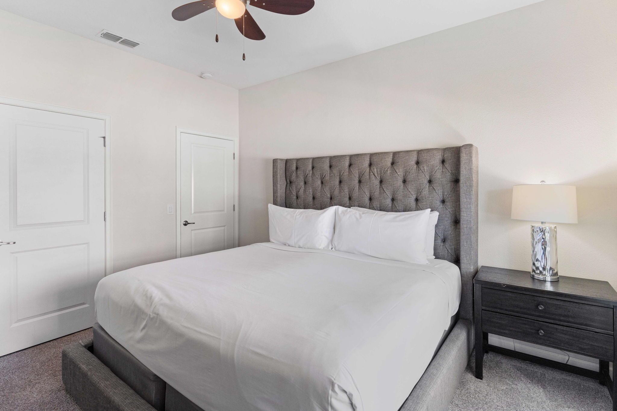 A neatly arranged bedroom with a large bed, a bedside table with a lamp, and a ceiling fan.
